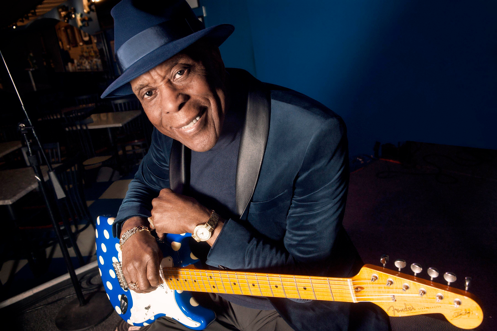 Buddy Guy looks into the camera holding a blue poka-dotted guitar and smiling.