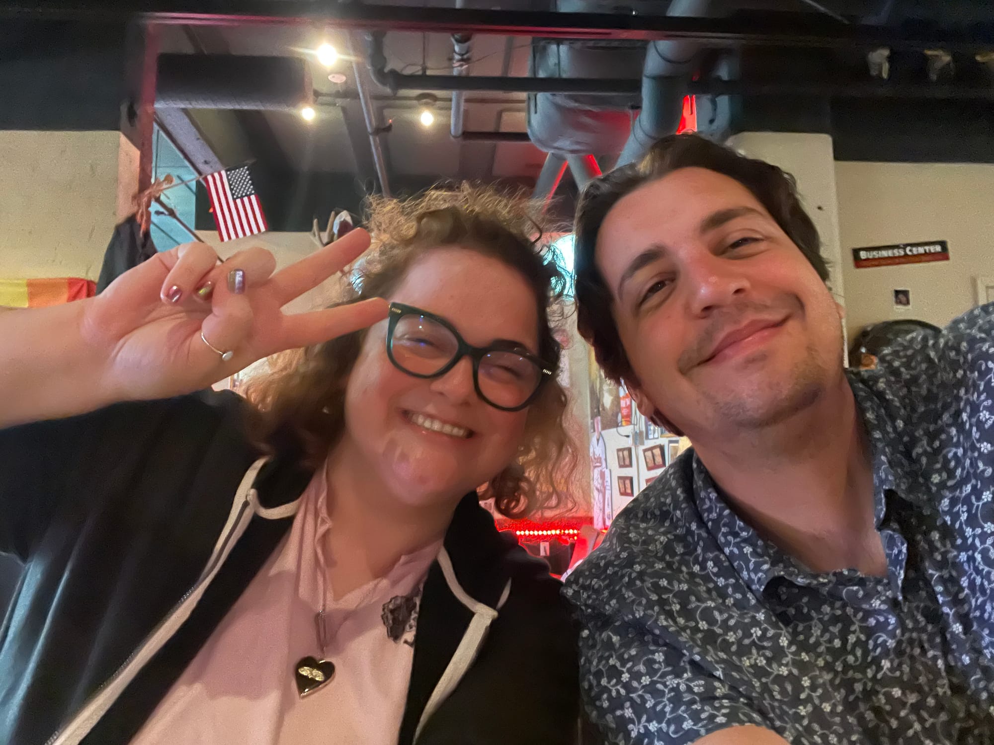 A picture of two people smiling into a camera, the woman on the left makes a peace sign and the man on the right leans into her smiling. They appear to be in a loosely industrial setting.